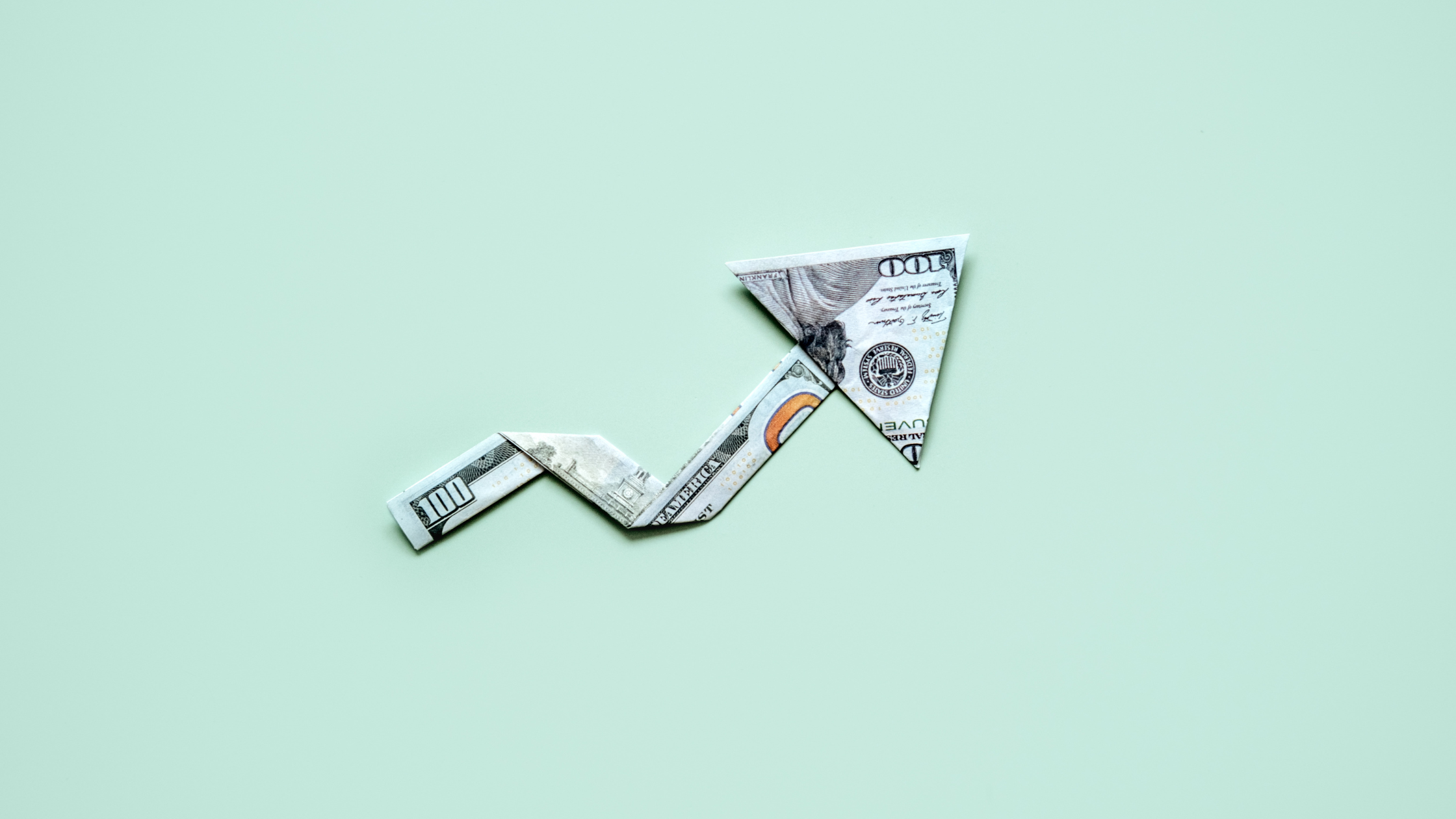 A one-hundred-dollar bill folded into the shape of an upward-trending arrow, symbolizing financial growth or investment gains, set against a minimalistic light green background.