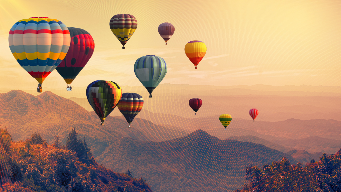 A dozen colorful hot-air balloons drift above layered, forested mountains in warm golden sunrise light.